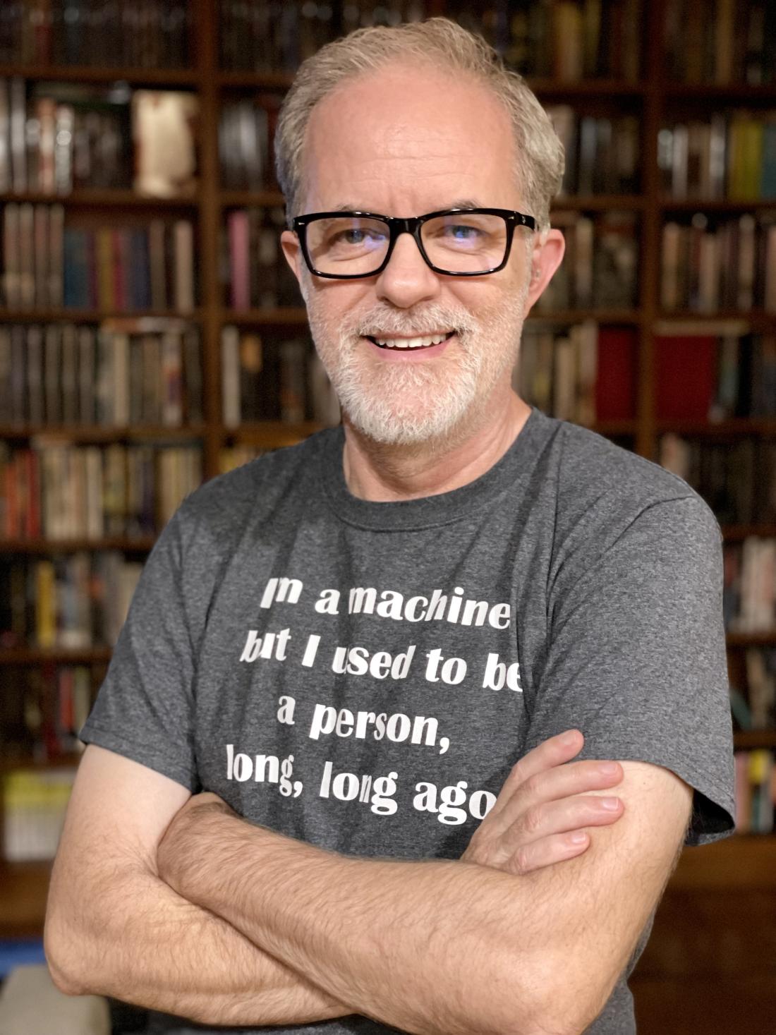 man with glasses standing in front of a bookshelf smiling into camera